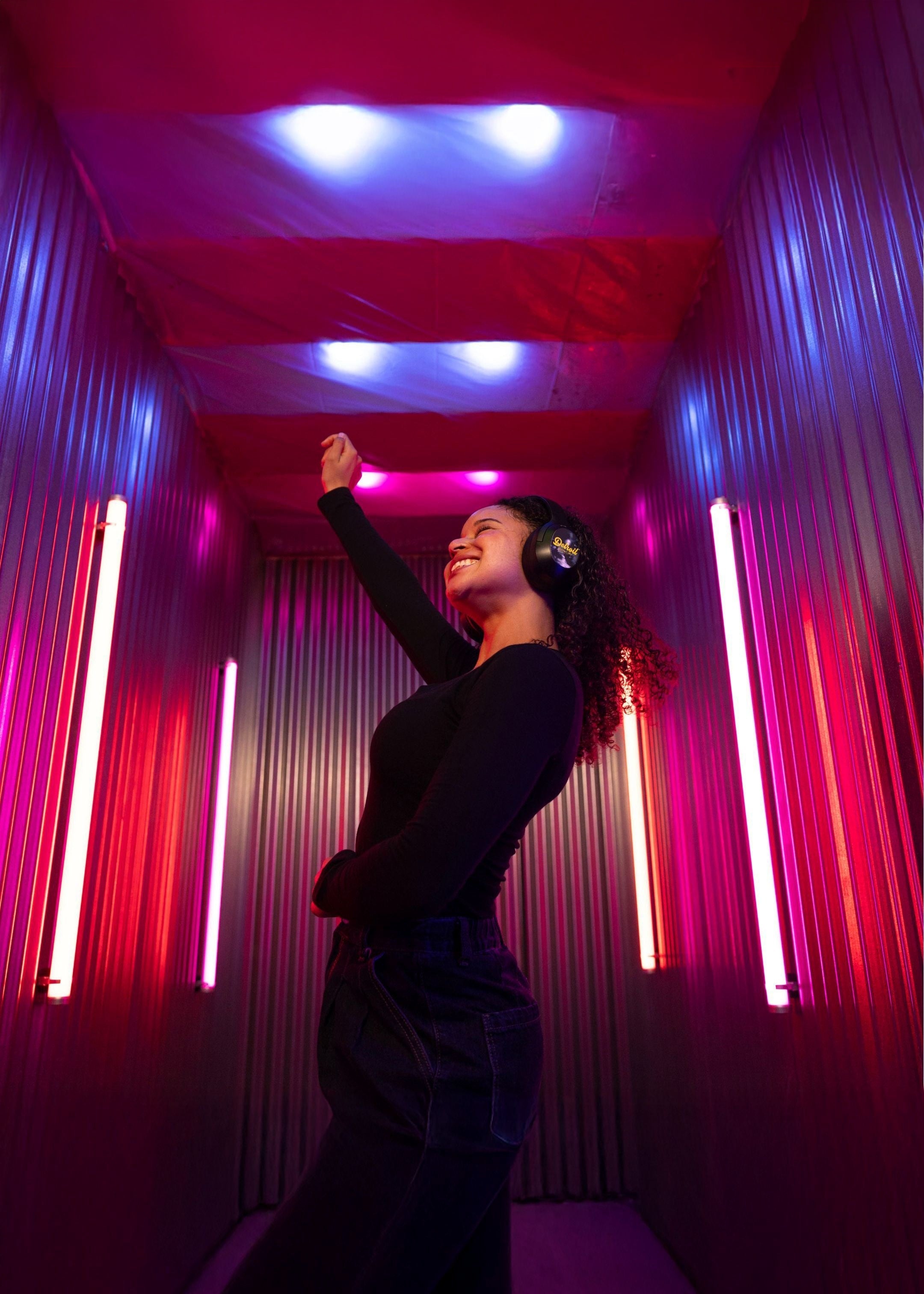 A woman wearing black headphones and a black long-sleeve top stands in a narrow corridor with pink, purple, and blue neon lights, looking upward and smiling.